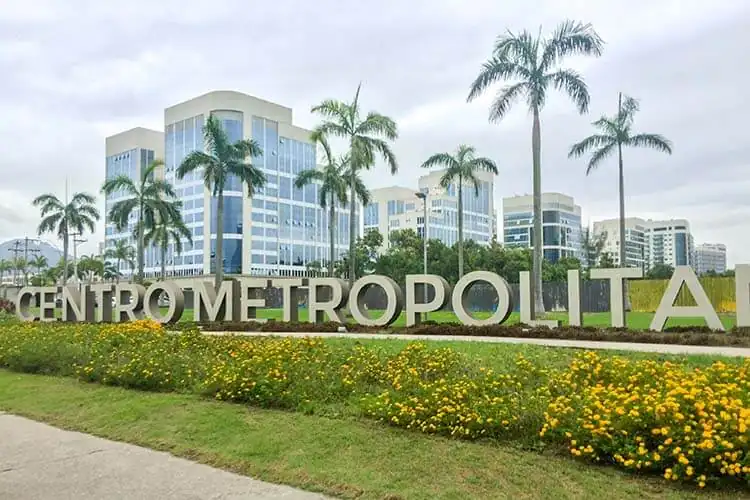 Letreiro do Centro Metropolitano, na Barra da Tijuca, com prédios comerciais, palmeiras e canteiro de flores amarelas